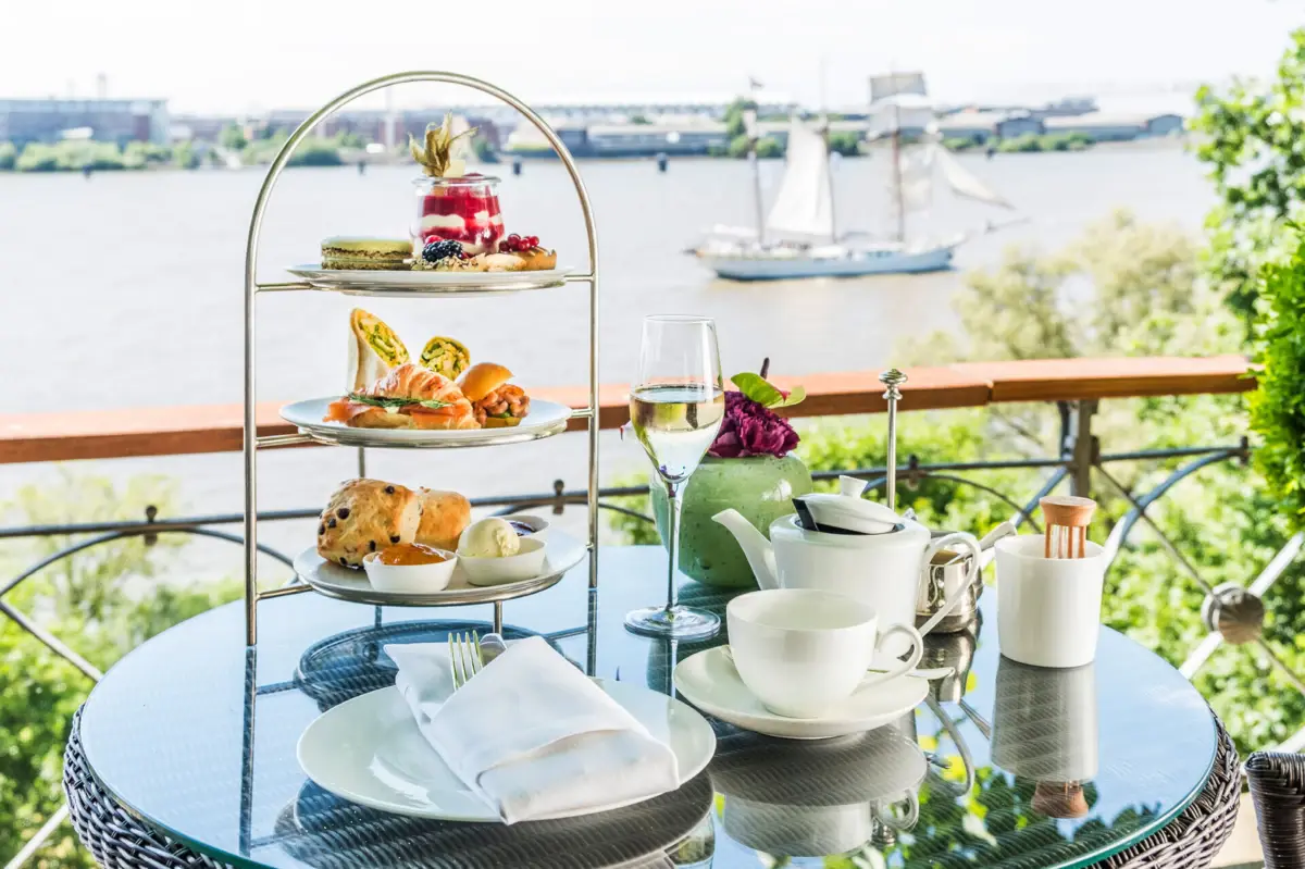Englische Teezeit mit herzhaften und süßen Kleinigkeiten serviert auf einem Tisch auf der Lindenterrasse des Hotels Louis C. Jacob mit Blick auf die Elbe und ein Segelboot.