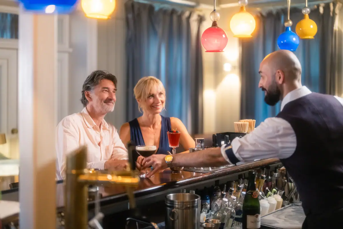 LCJ_egr_Sommer23_Bar_1611.jpg Ein Paar sitzt lächelnd am Tresen der Jacobs Bar und wird von einem Barkeeper bedient.