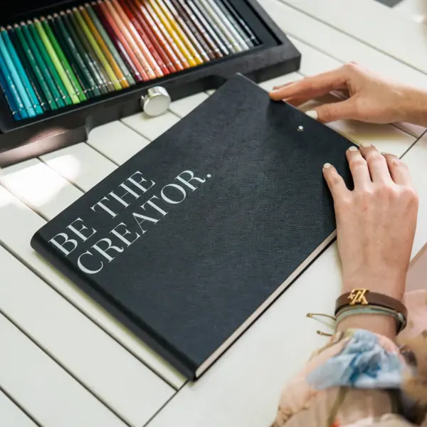 Hände einer Person auf einem Buch mit Buntstiften im Hintergrund.