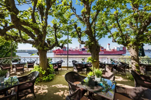 A group of tables and chairs under trees by a body of water.