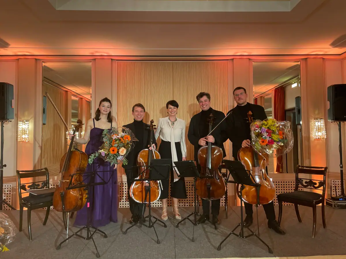 Vier Musiker des TONALI Celloquartetts stehen zusammen mit der Hoteldirektorin Judith Fuchs-Eckhoff mit ihren Celli vor einer Wand in einem Saal des Hotels Louis C. Jacob.