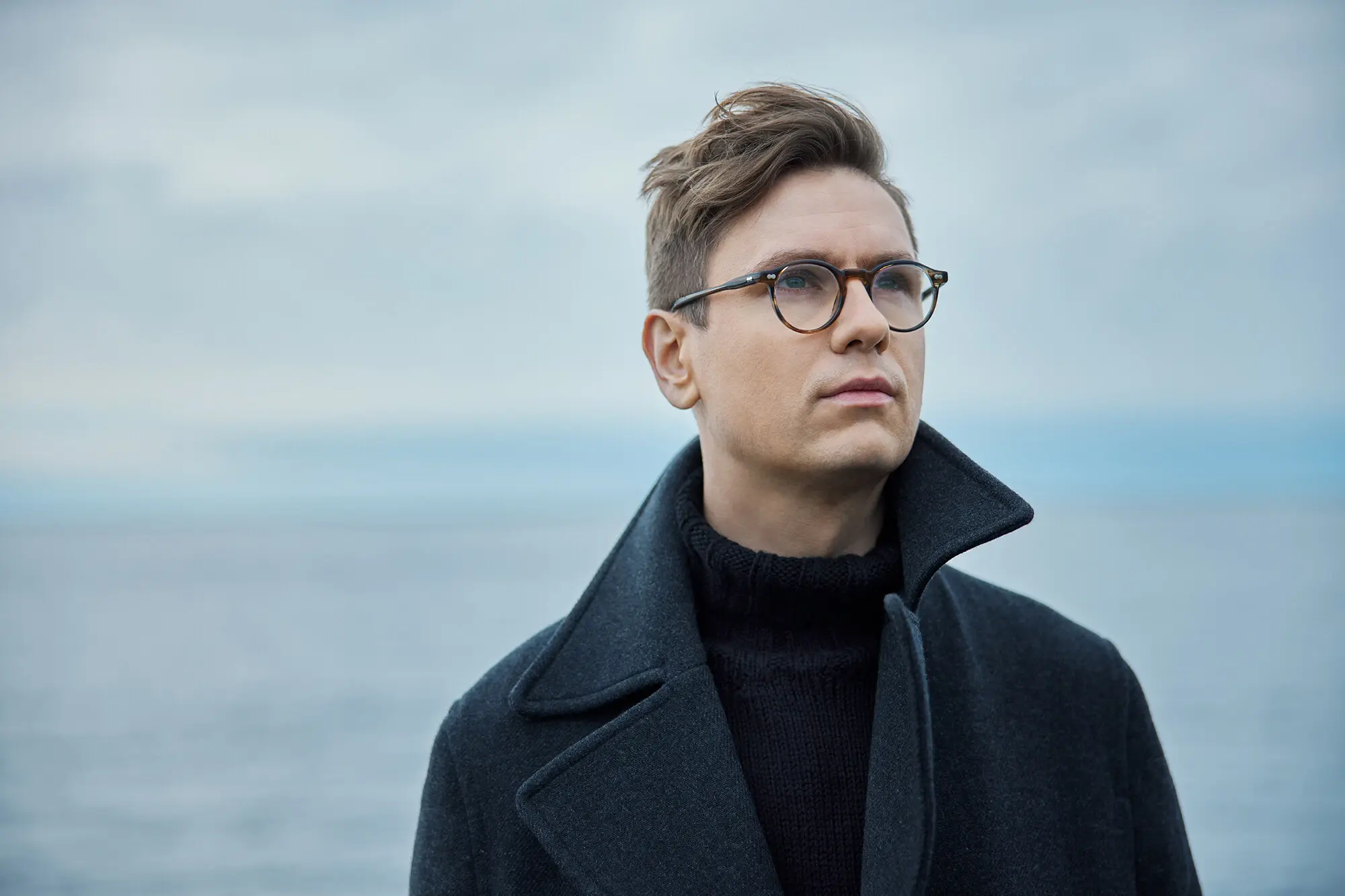 Víkingur Ólafsson Ein Mann in Mantel und mit Brille steht am Wasser und schaut in die Ferne.