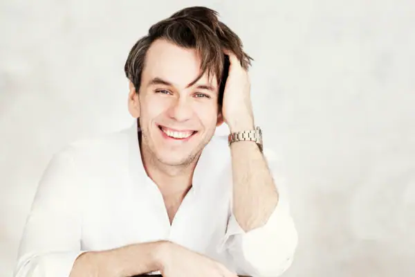 Benjamin Bernheim Eine Mann in weißem Hemd sitzt vor einem hellen Hintergrund und lacht in die Kamera.