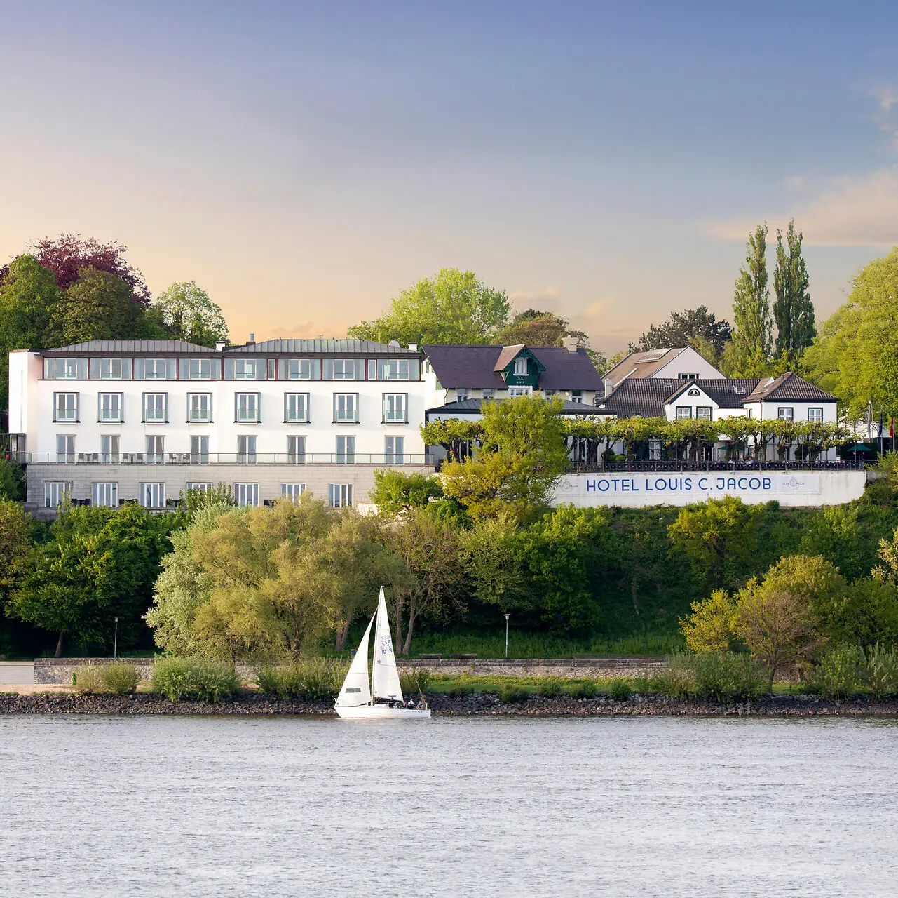 Das Louis C. Jacob Hotel an der Elbe mit einem Segelboot davor im Wasser.