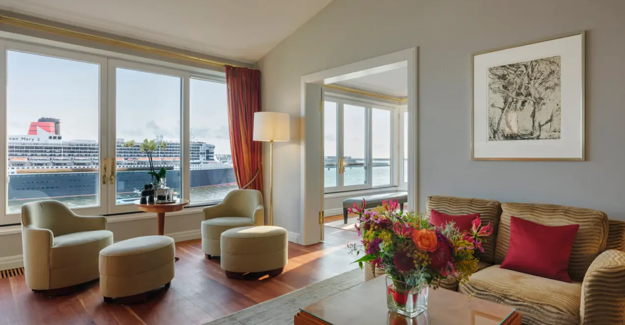 Living area of the Elbe suite in the Hotel Louis C. Jacob with couch, armchairs and coffee table in front of a large window with a view of the Elbe.