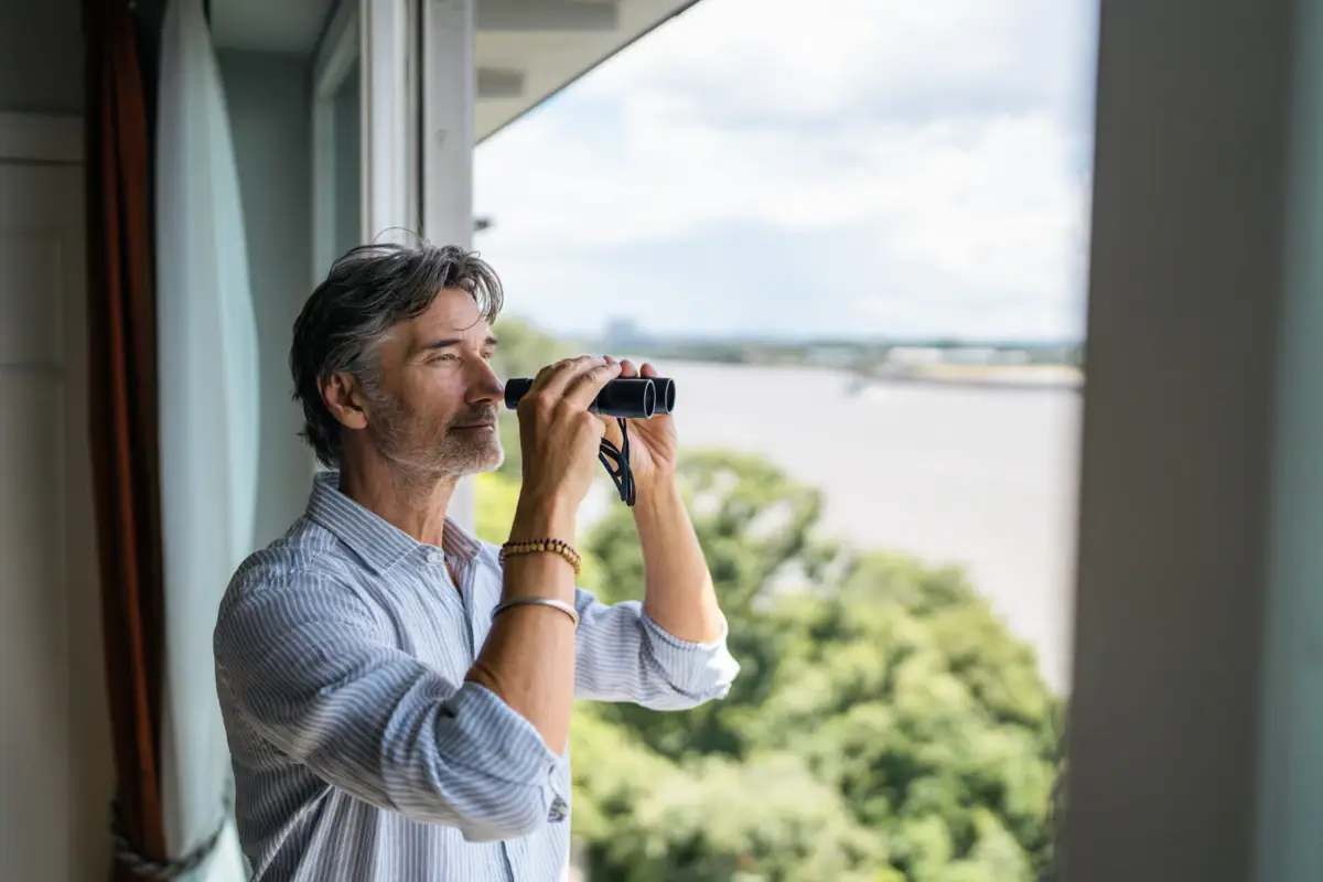 Ein Mann schaut durch ein Fernglas aus einem Zimmer vom Louis C. Jacob Hotel Hamburg auf die Elbe.