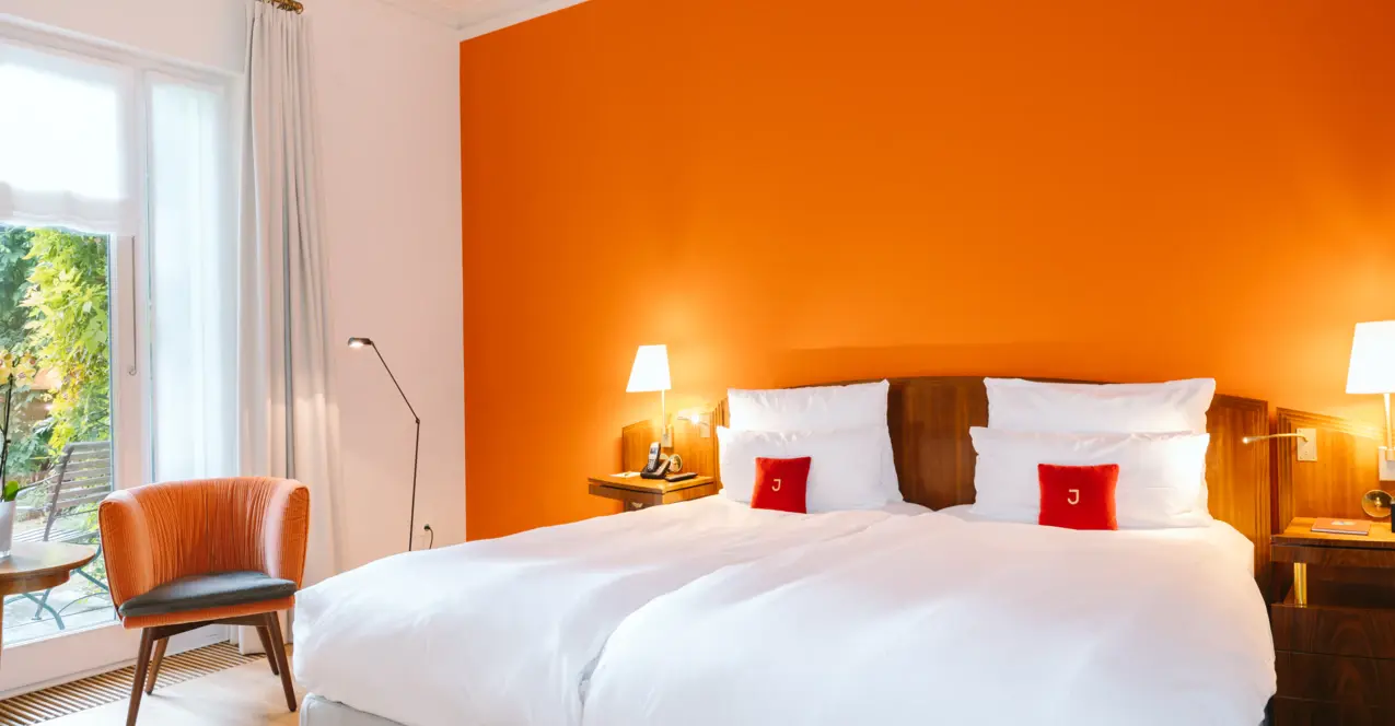 Bright hotel room with two beds, orange accent wall, an armchair, two bedside tables with lamps and a large window overlooking a garden.