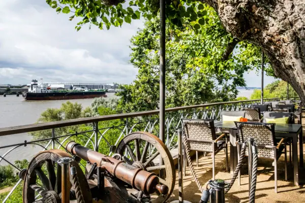 Lindenterrasse des Hotels Louis C. Jacob mit Gartenstühlen und Tischen sowie einer kleinen Kanone. Von der Reling kann man die Elbe und ein Containerschiff sehen.