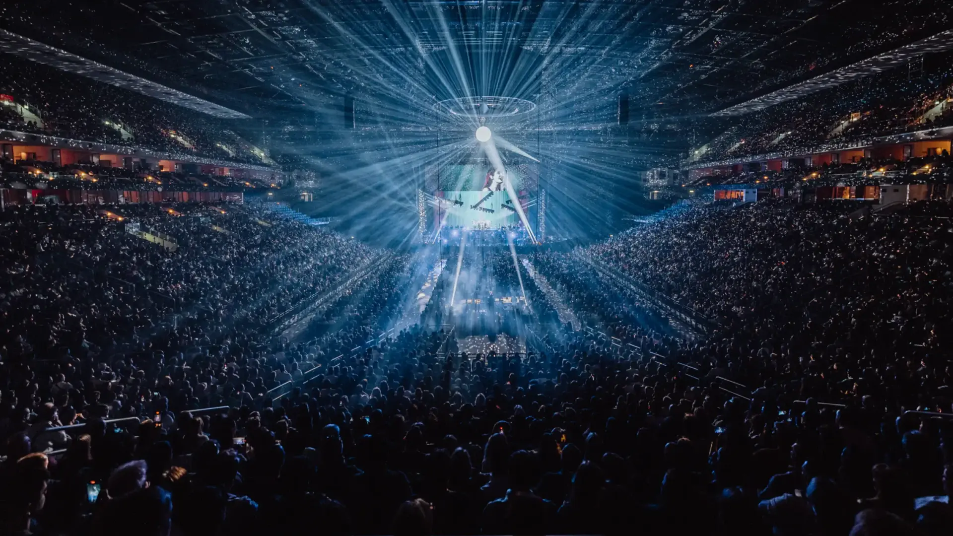 A large arena filled with people during a concert. The room is dark and blue beams of light shine through the air.