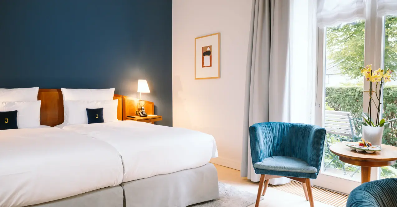 Stylishly furnished hotel room with blue accent wall, cozy double bed, elegant armchairs and a view of the greenery through floor-to-ceiling windows.