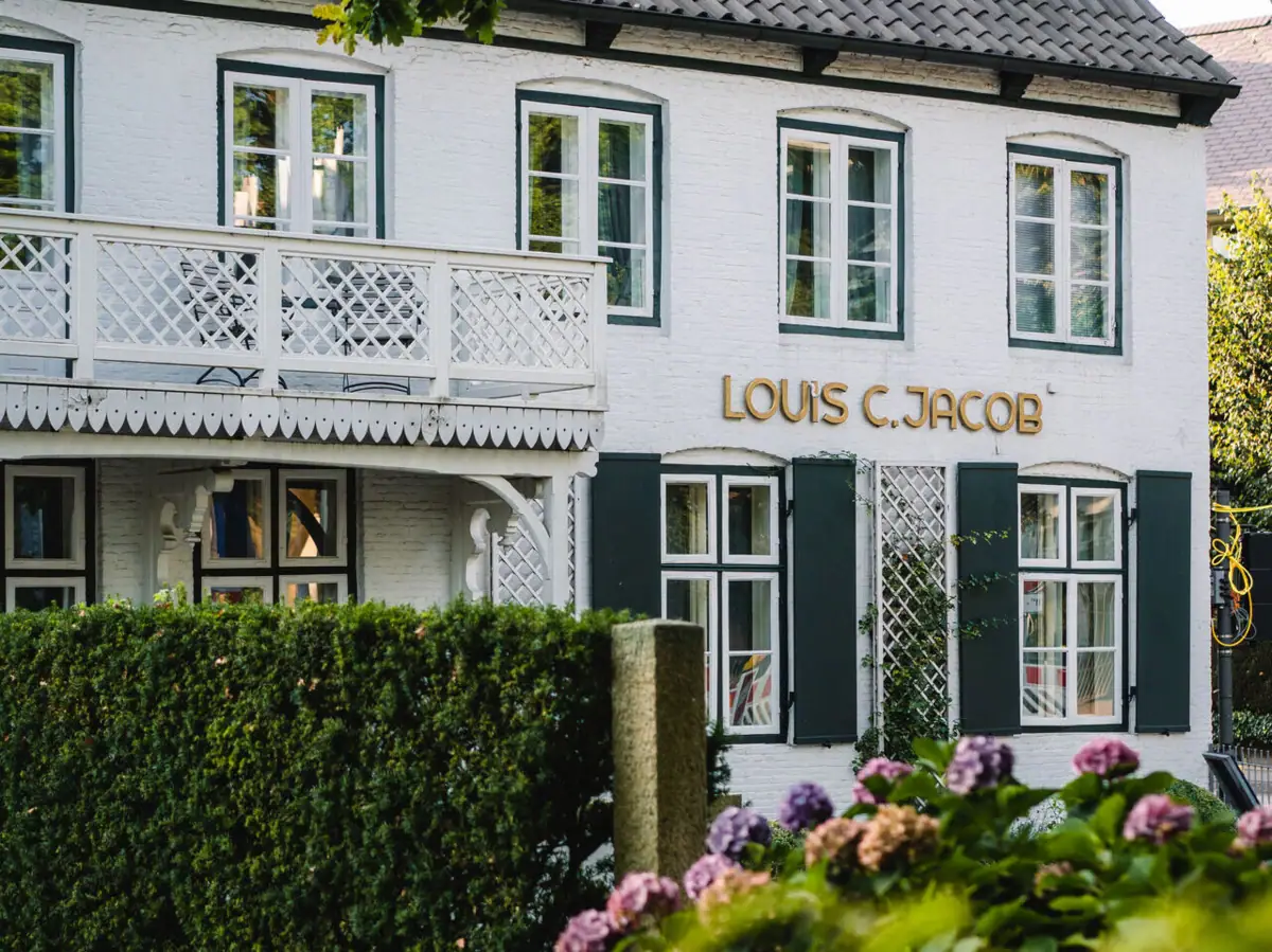 Das Louis C. Jacob mit weißem Balkon und einer grünen Hecke davor.