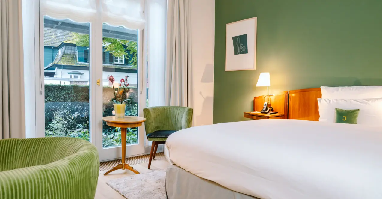 Stylishly furnished hotel room with green accent wall, cozy double bed, elegant armchairs and a view of the greenery through floor-to-ceiling windows.
