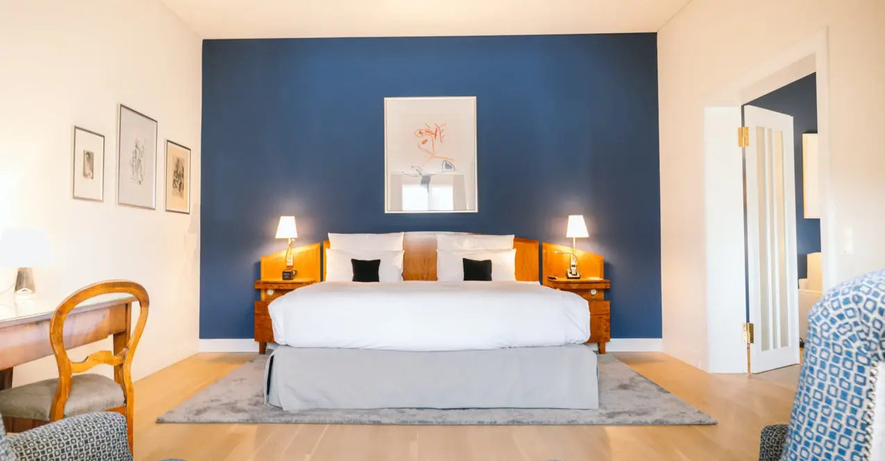 A hotel room at the Louis C. Jacob with a bed and a blue wall, equipped with pillows, bed linen and a bedside lamp.