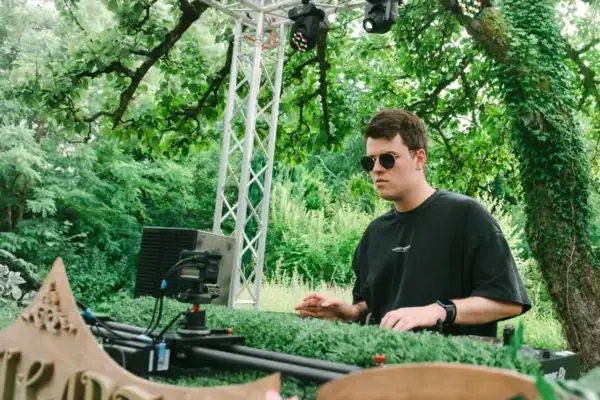 Marten Toneyard Ein Mann mit Sonnenbrille legt als DJ im Freien vor grünen Bäumen auf.