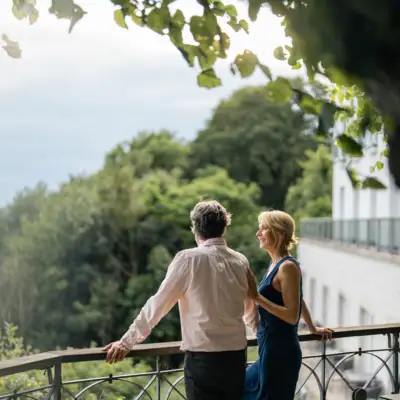 A man and woman standing on a balcony with trees and sky in the background.