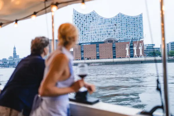 Ein Mann und eine Frau stehen an Bord der Barkasse MS Jacob und schauen auf die Elbe und die Elbphilharmonie.