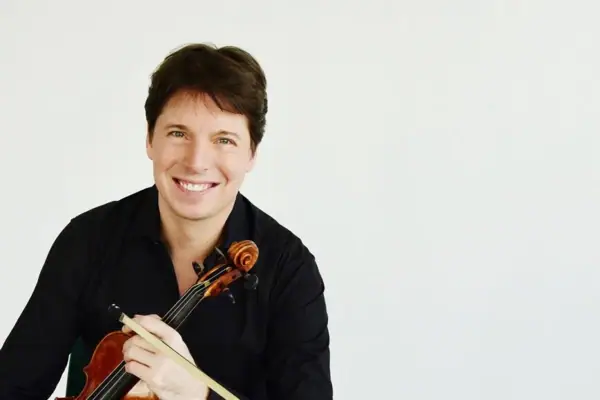 Joshua Bell A man is sitting in front of a white background, smiling and holding a violin.