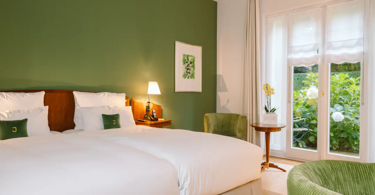 Stylishly furnished hotel room with green accent wall, cozy double bed, elegant armchairs and a view of the greenery through floor-to-ceiling windows.