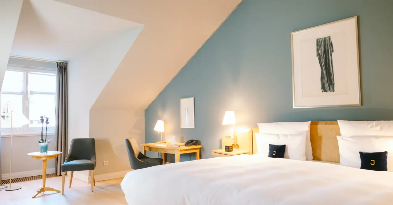 Brightly furnished hotel room with sloping ceiling, large double bed in front of blue-grey wall, two bedside tables with lamps and seating area with table and chairs by the window.