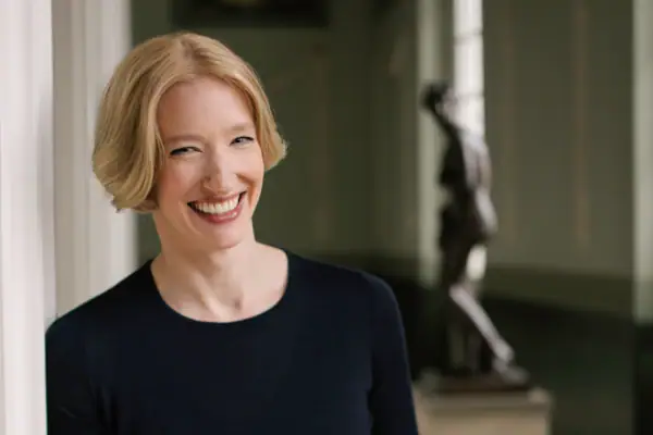 Eine Frau mit kurzen blonden Haaren steht an eine Säule gelehnt und lächelt in die Kamera.