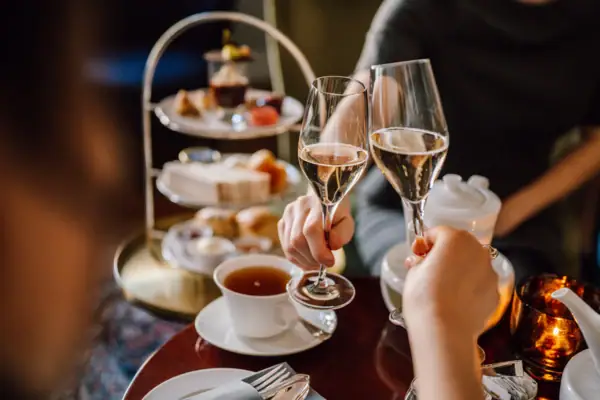 Afternoon tea at the Hotel Louis C. Jacob - a teacup is on the table and two people are toasting with champagne. In the background you can see an étagère with scones, sandwiches and sweet desserts.