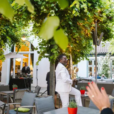 A man in a white suit is playing the saxophone under a tree.